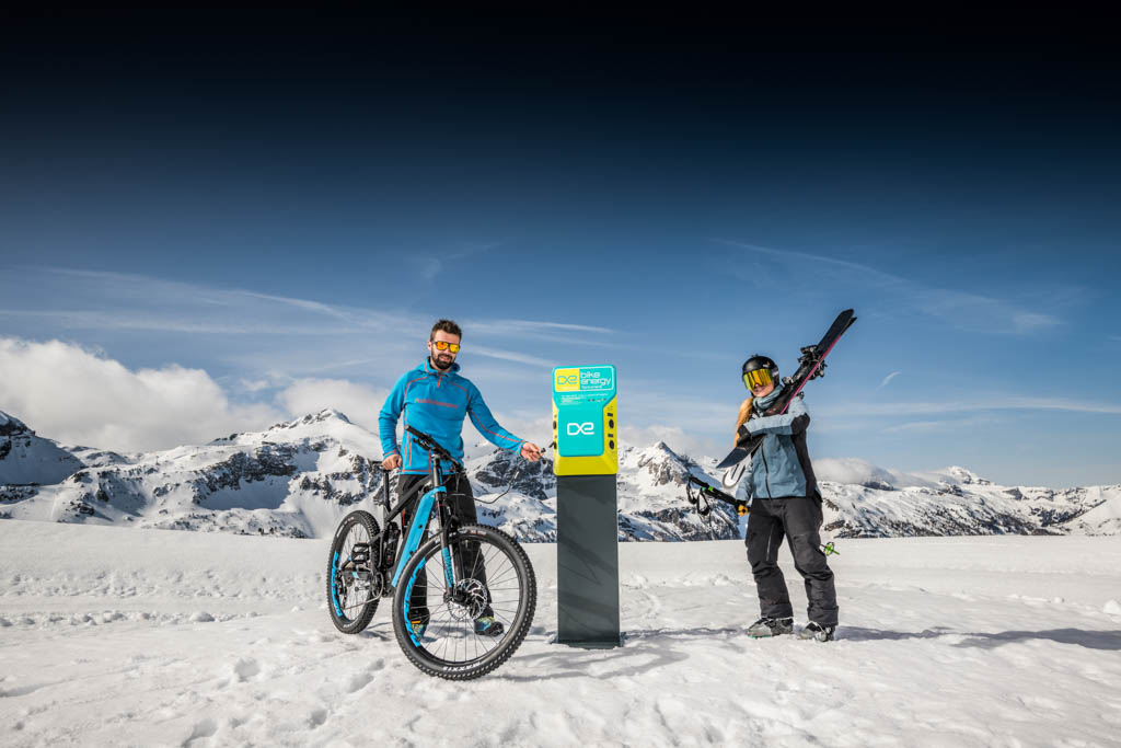 Herr lädt sein E-Bike an der Ladestation am Berg auf, Dame trägt die Schi auf den Schultern